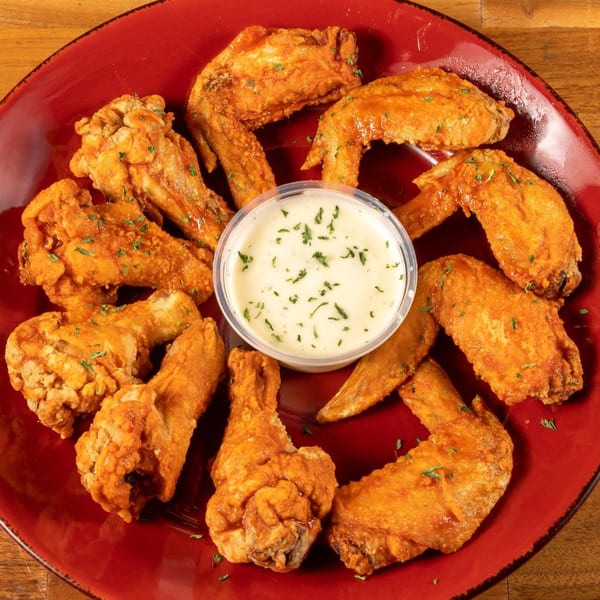 A red plate filled with golden-brown chicken wings, some sauced, surrounding a cup of ranch dressing.